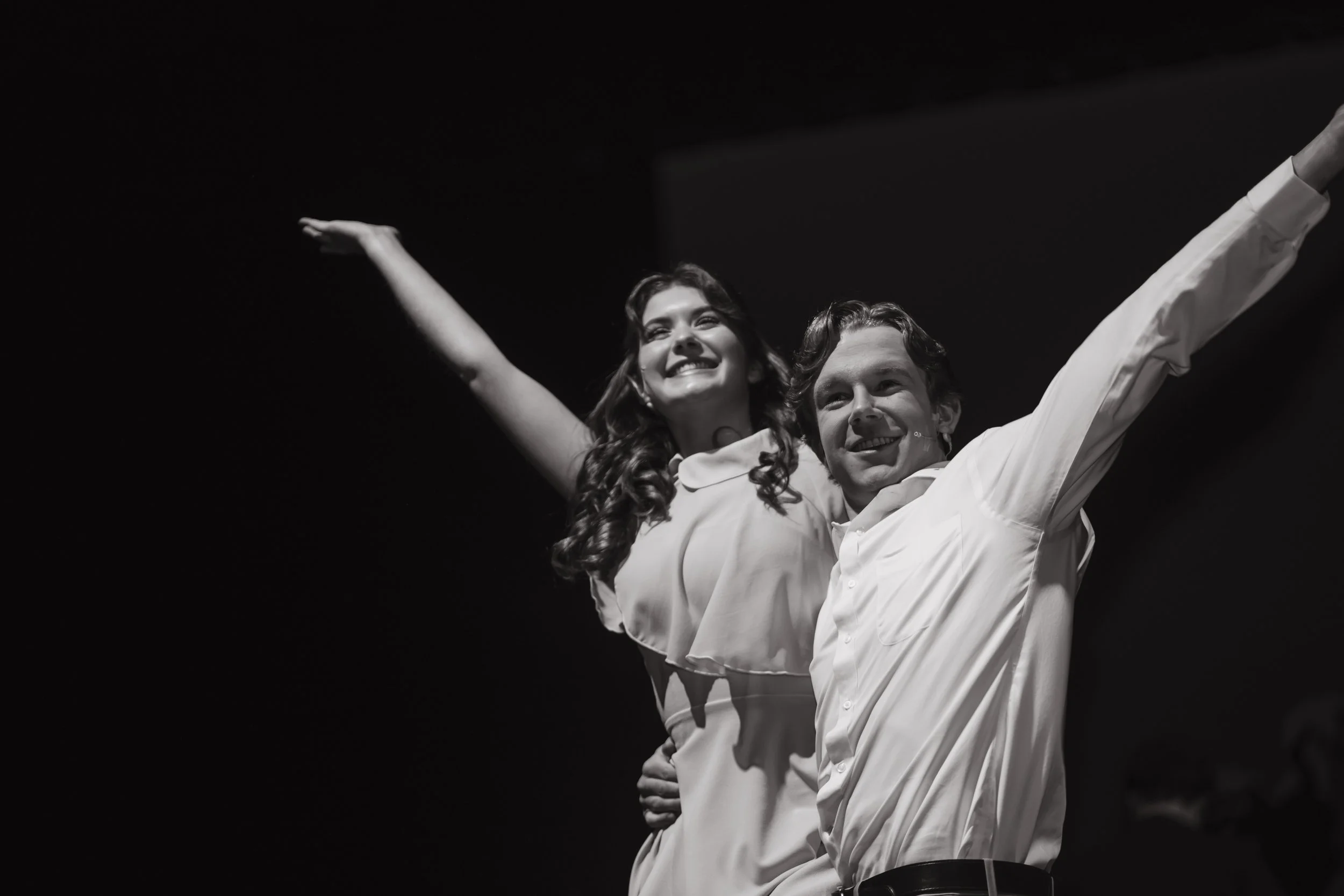 Black and white photo of a smiling woman with her arms raised being held up by a man with his arms also raised, both looking cheerful and celebrating.