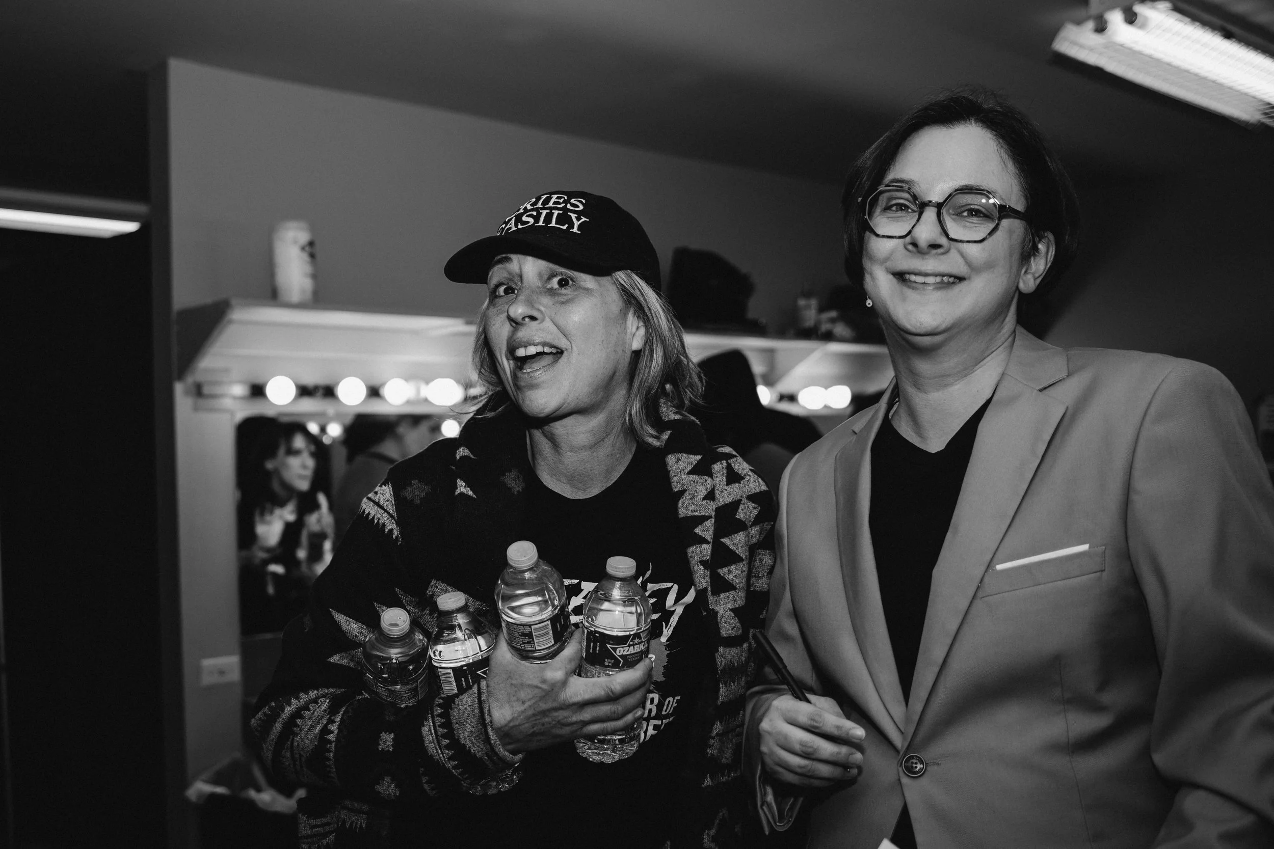 Two women smiling at an indoor event, one is holding four water bottles and wearing a baseball cap, the other is wearing glasses and a blazer.