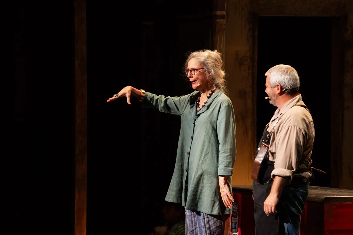 An older woman with gray hair, glasses, and a necklace, engaged in a conversation or performance, gesturing with her arm extended towards the side, while a man with gray hair and a beard, holding a guitar, listens attentively.
