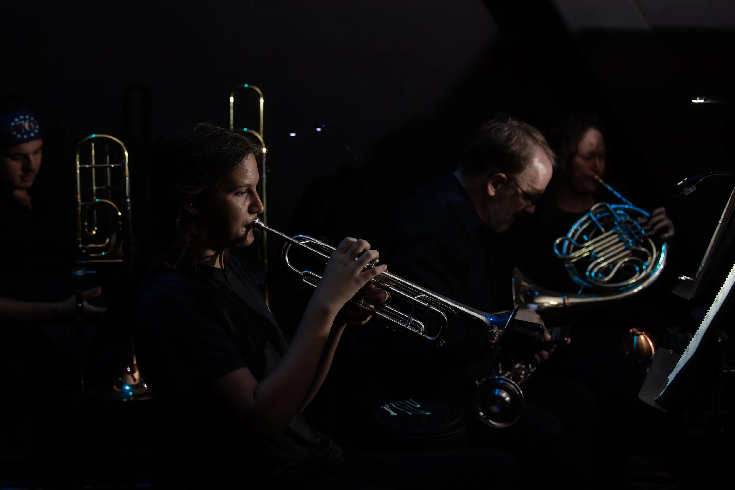 Musicians playing brass instruments in a dark setting, with one woman in focus playing a trumpet.