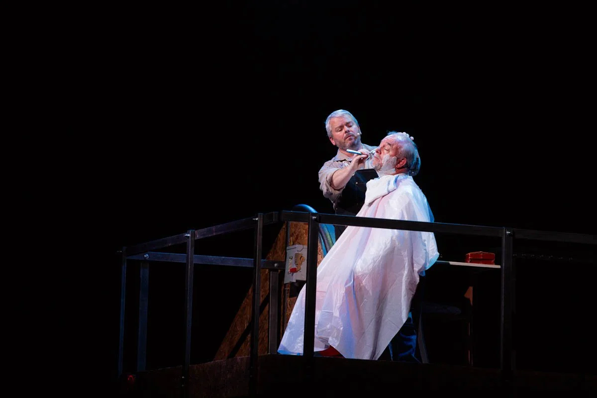 A man with grey hair and a beard receiving a haircut from a barber on a stage with a black background. The man is seated, covered with a white cape, while the barber uses scissors and a comb.