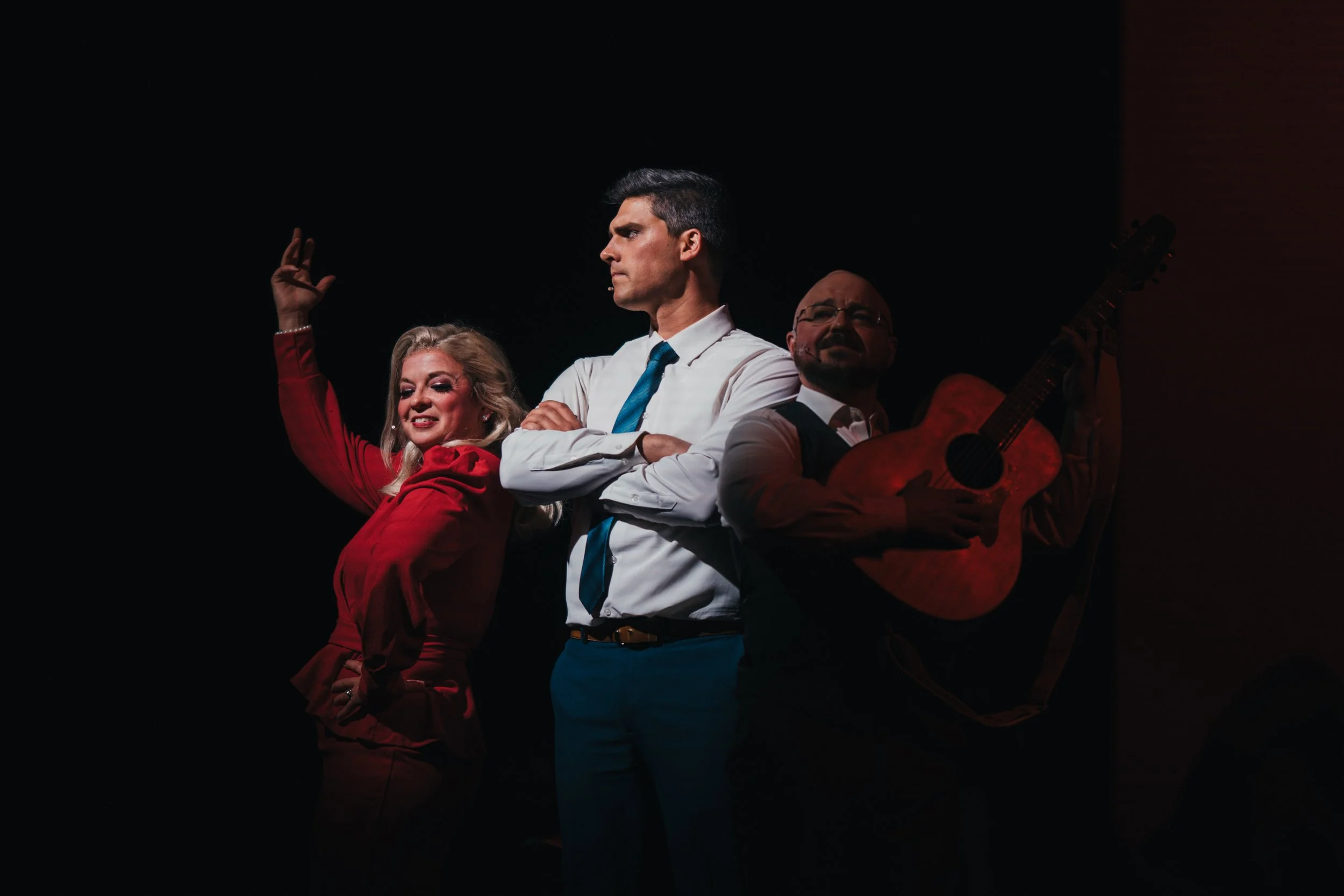 Three performers on stage: a woman in red with blonde hair, a man in a white shirt and blue tie with dark hair, and a man in a vest holding a guitar, with a dark background.