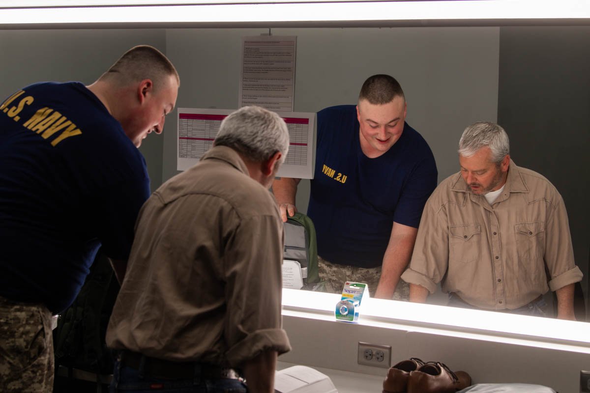 Four men, including two in U.S. Navy uniforms, look into a mirror in a locker room or dressing area, with fluorescent lighting and a small box of eyeshadow or makeup on the counter.