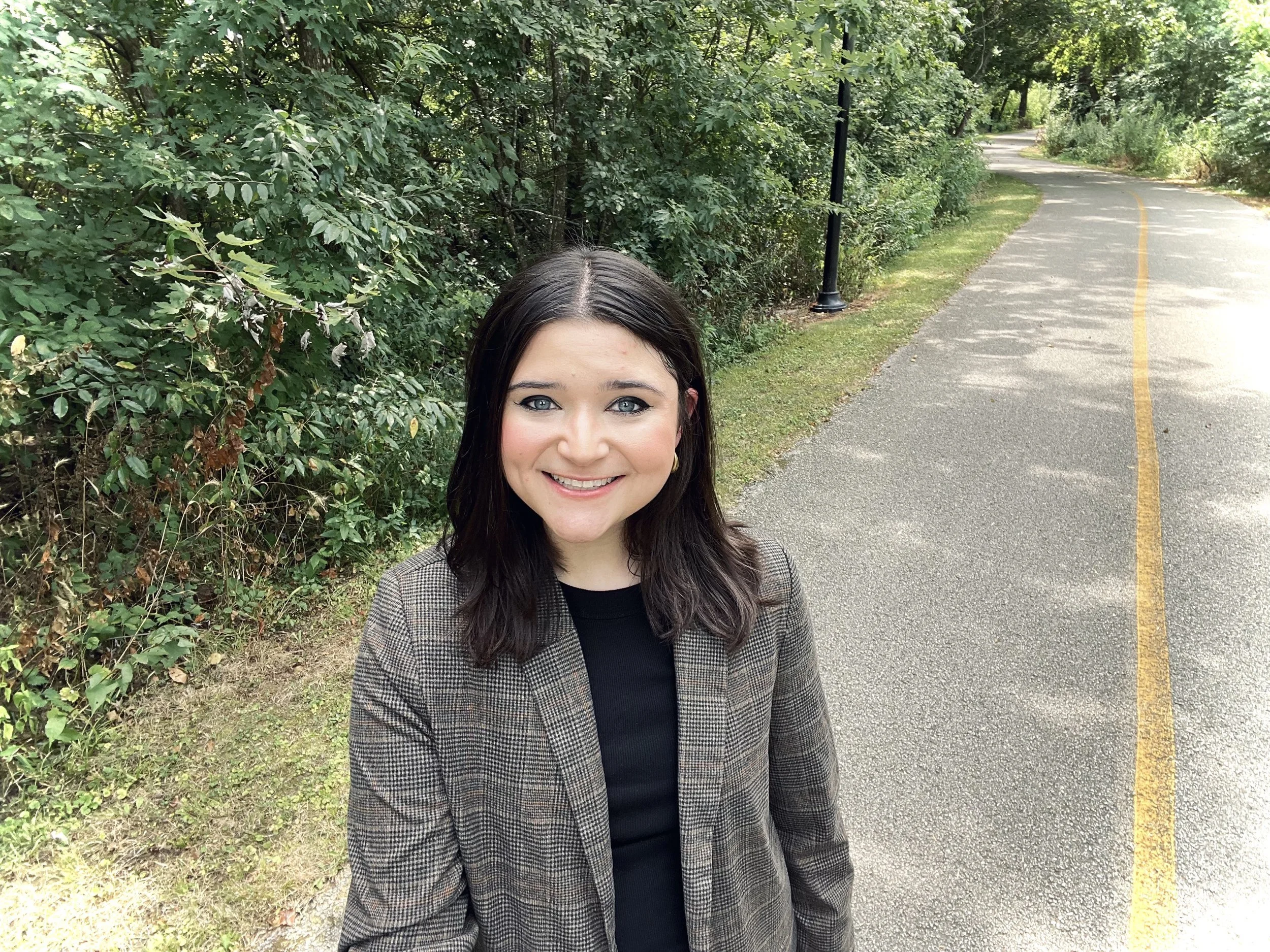 A young woman smiling outdoors on a paved path with green bushes and trees in the background.