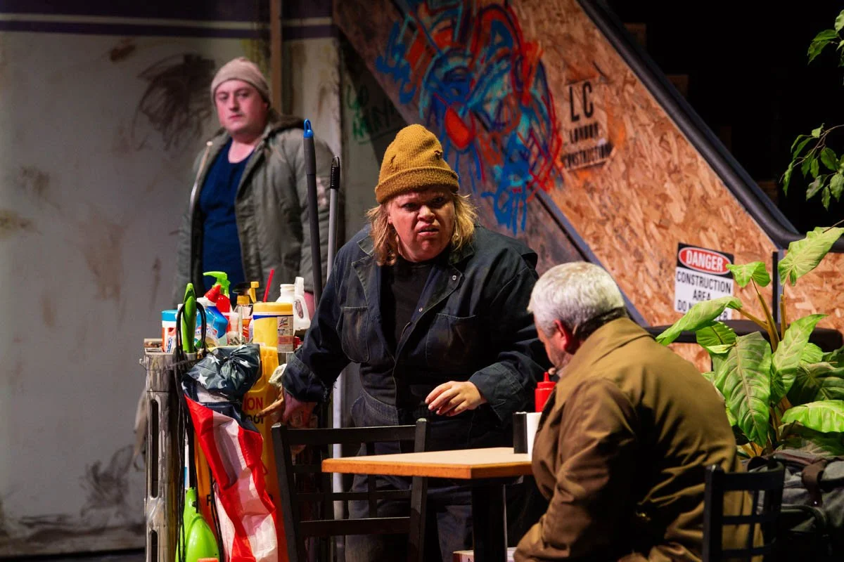 Three people in a restaurant or cafe, with one person standing behind a counter with cleaning supplies, another person sitting at a table with a gray hair, and a woman in a yellow beanie talking to her. There is graffiti art on the wall in the backgr