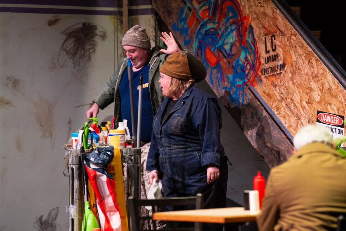 Two men on a stage with a cart full of cleaning supplies, one pointing and the other speaking, with a wooden wall decorated with graffiti art and safety signs in the background.