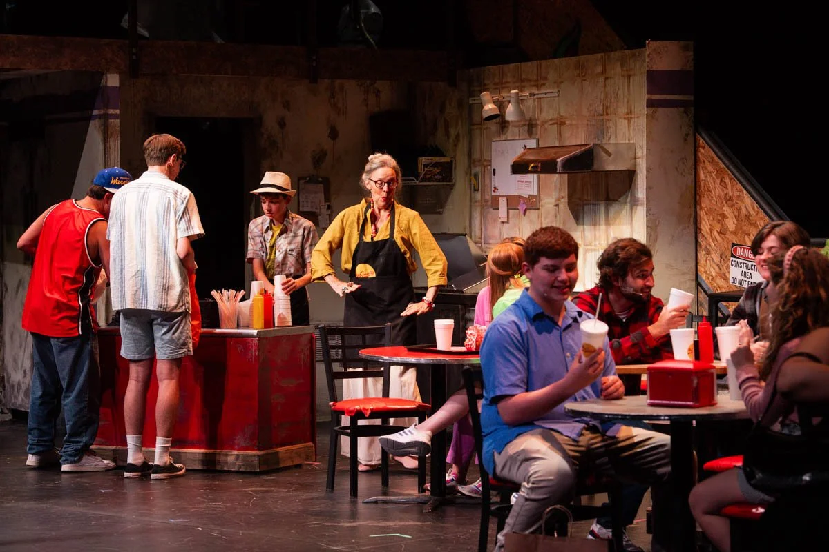 Actors on stage perform a scene set in a casual restaurant or diner, with people sitting at tables and others ordering food from a counter, in an indoor theatrical setting.