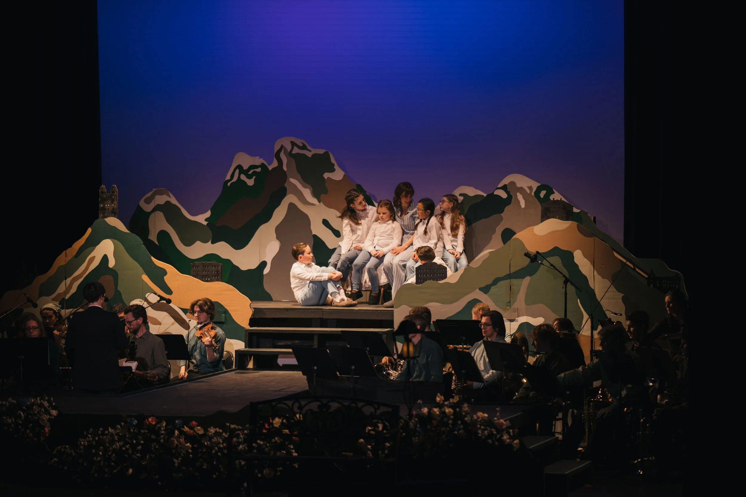 Children sitting on a stage with a mountain landscape backdrop, part of a theatrical performance, with musicians in the foreground.