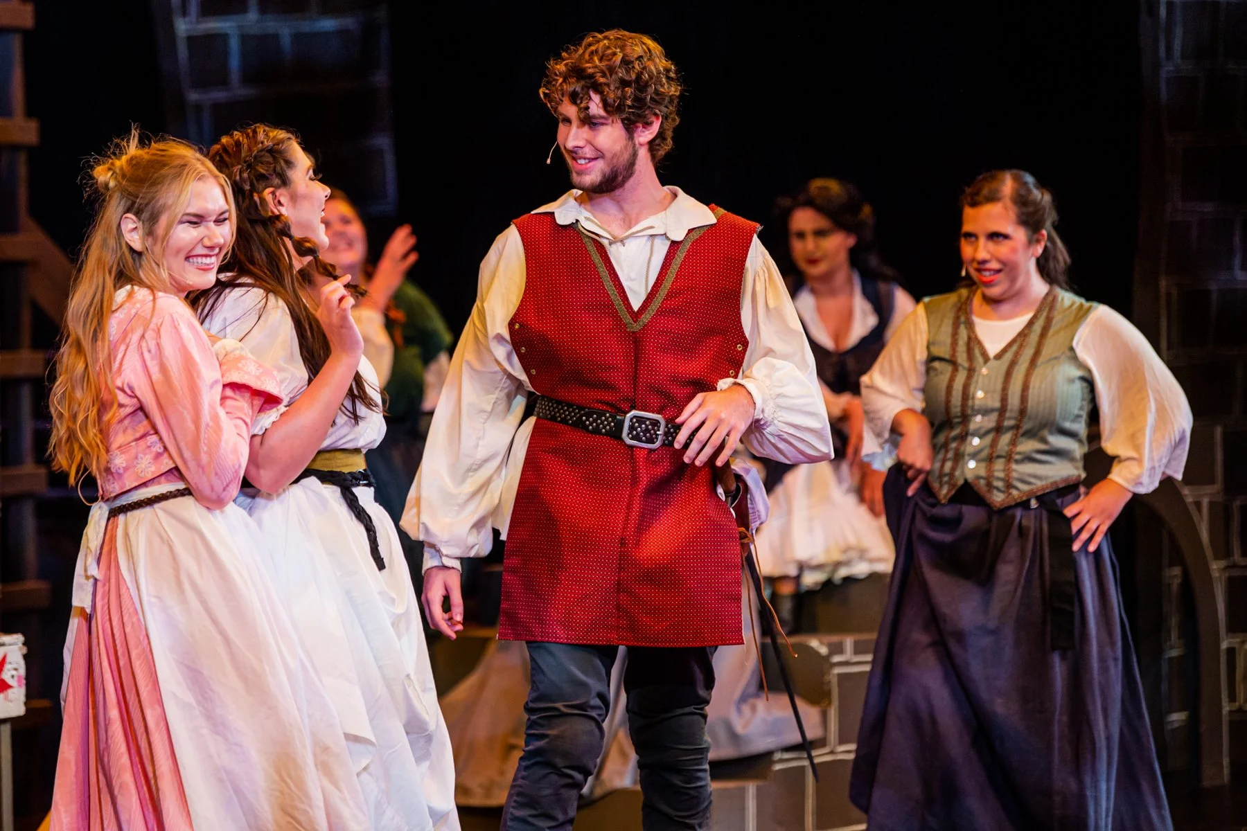 Cast of a theatrical play dressed in period costumes on stage, smiling and interacting with each other.
