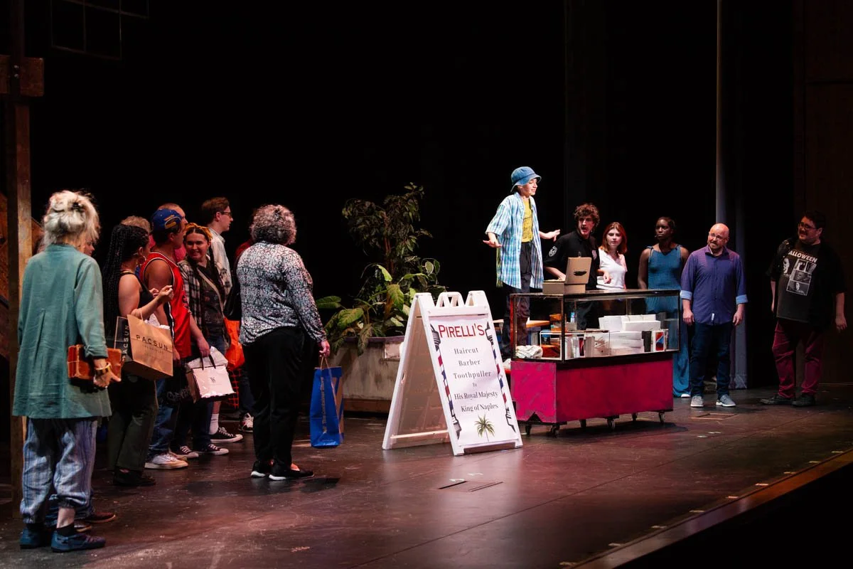 A group of people standing in line on a stage next to a person wearing a blue hat and plaid shirt, performing or presenting in front of the audience. There is a display case and a sign for PIRELLI'S on the stage.
