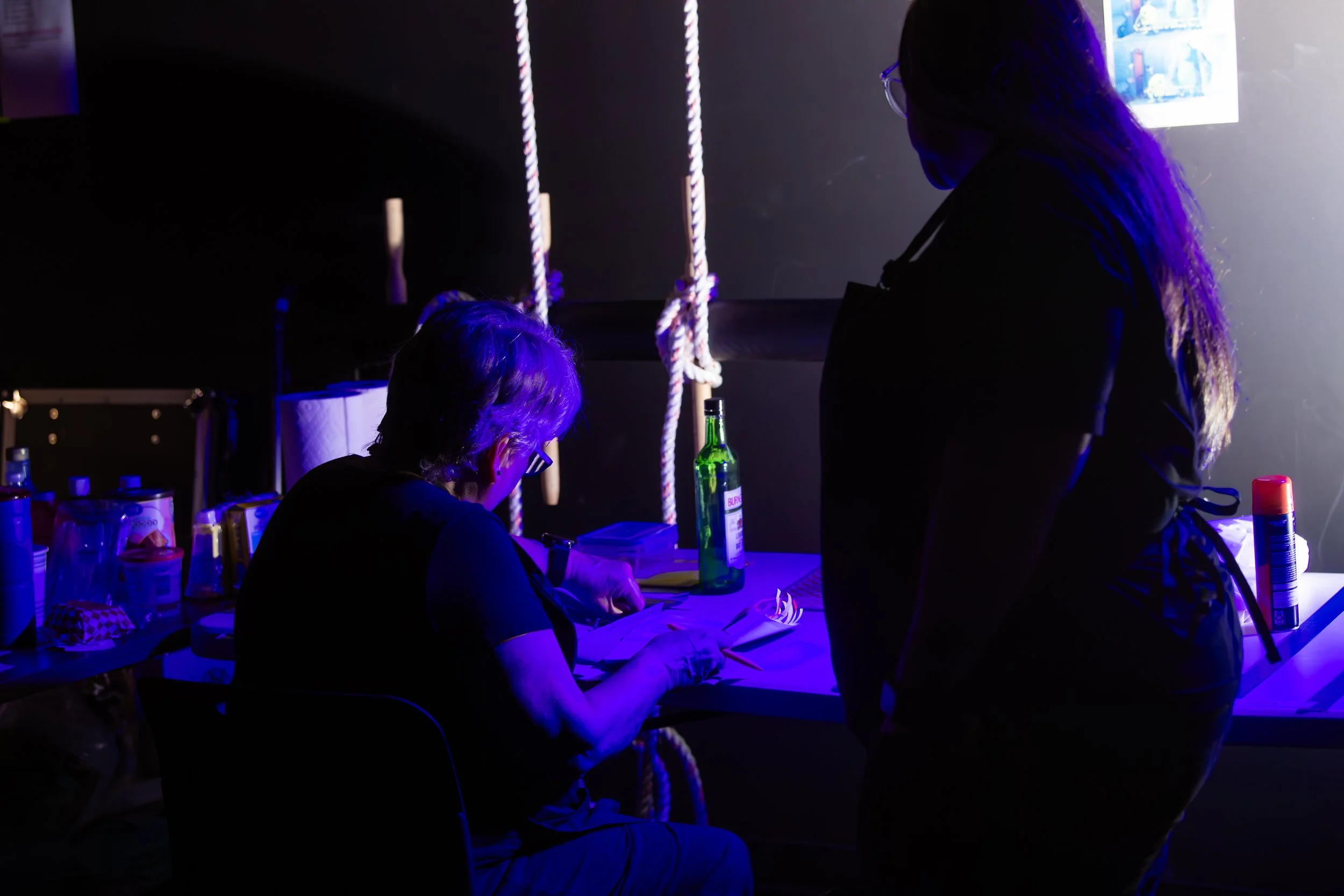 Two people working at a table in a dark room illuminated by purple light. One person is seated and writing, while the other stands beside them, observing. Items on the table include bottles, papers, and cups.