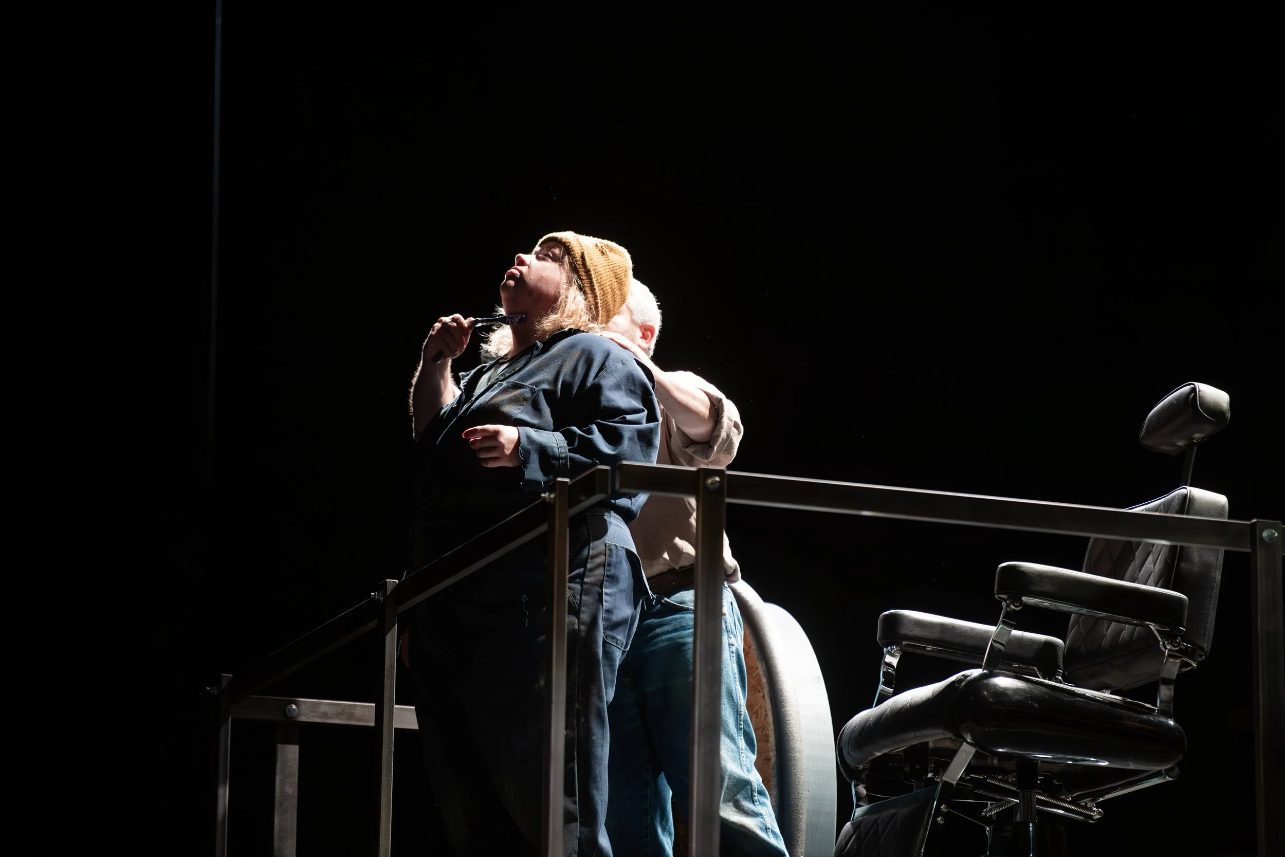 A woman with blonde hair wearing a brown beanie and dark jacket stands on a dimly lit stage, with an older person with white hair behind her, embracing her. There is a black office chair to the right and a ballet barre-like railing in front of them. 