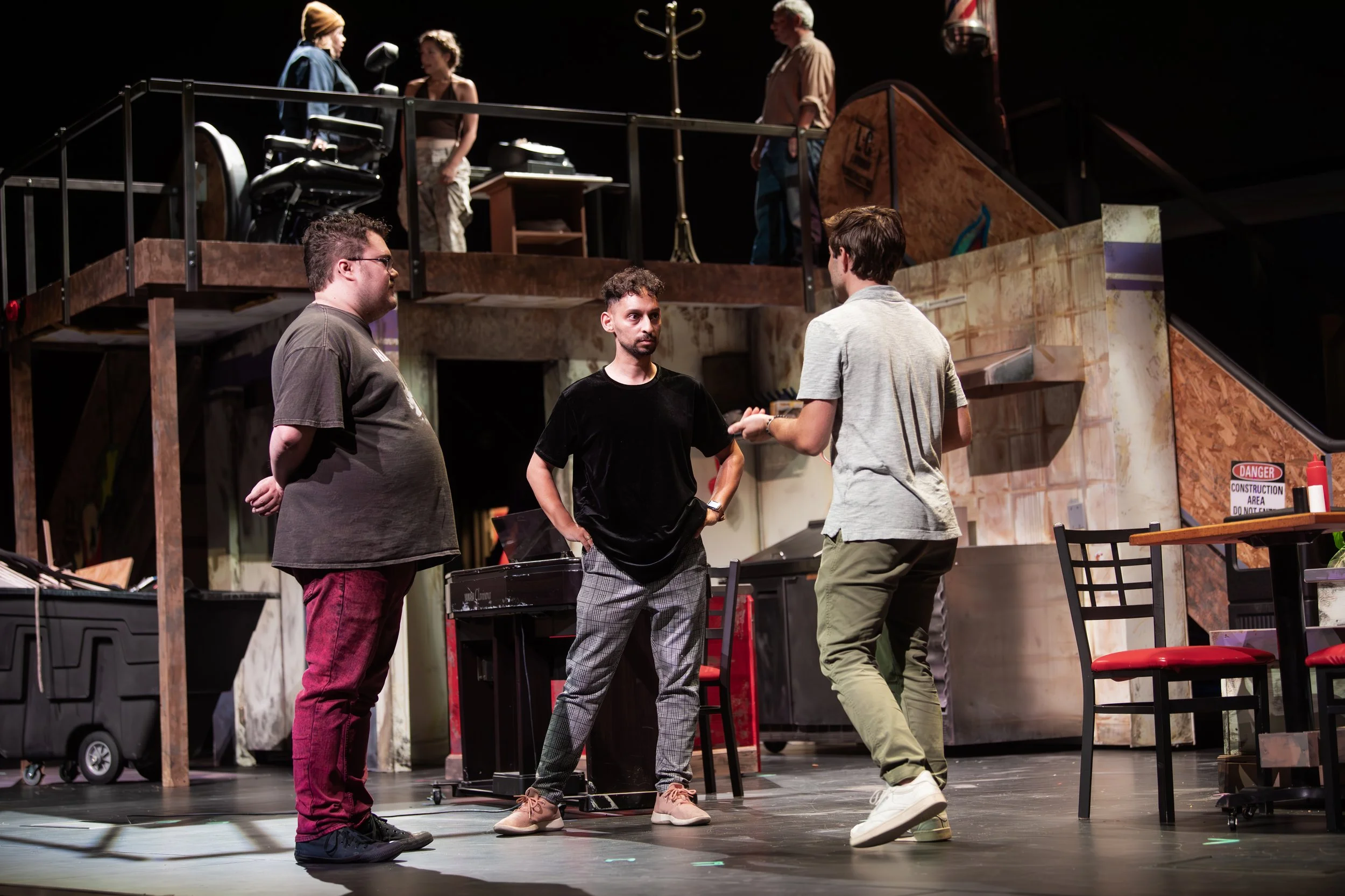 Three men having a conversation on stage during a theatrical rehearsal, with a two-level set and stage props in the background.