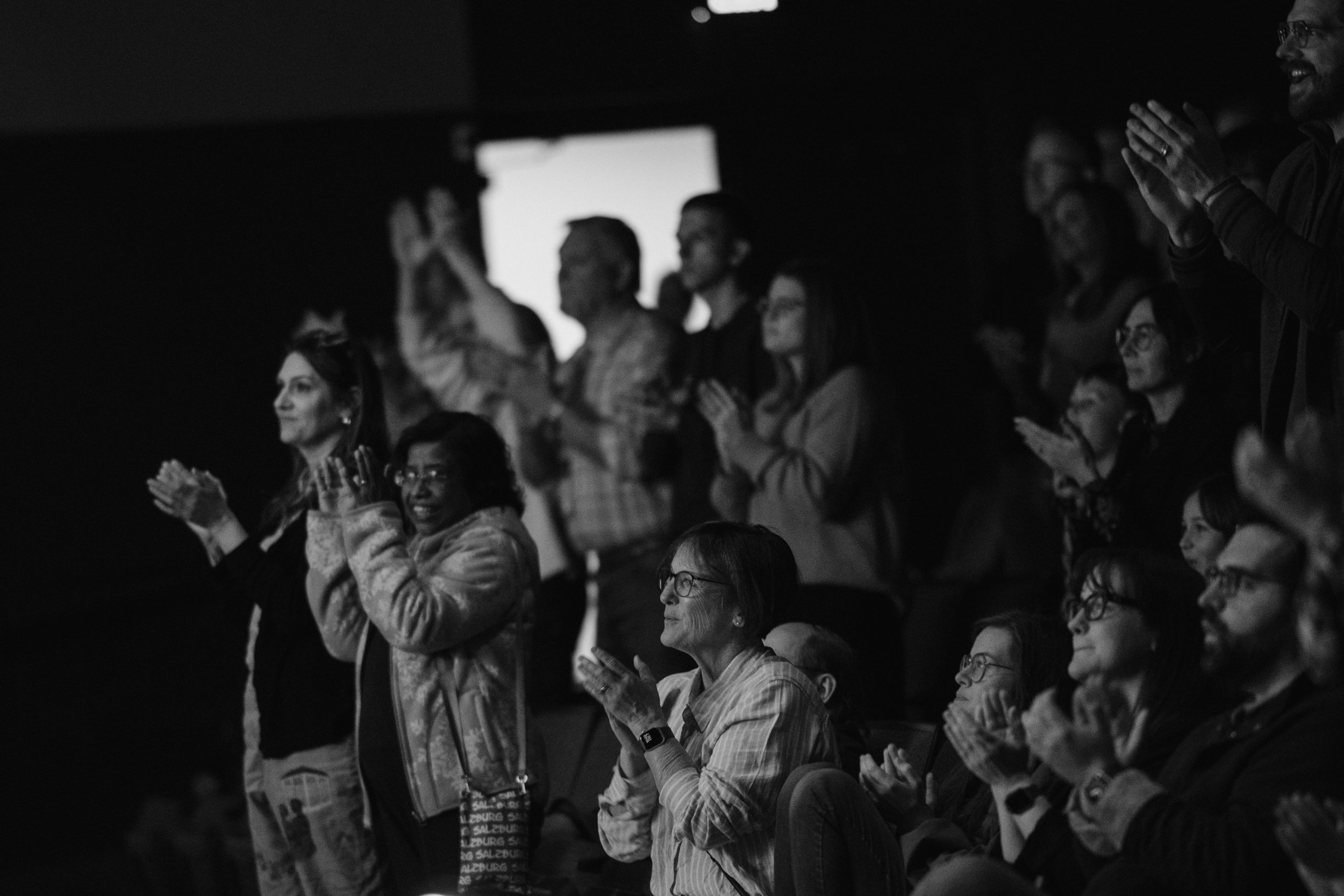 Audience members clapping and watching an event in a dark theater, some seated and others standing, with a large screen visible in the background.