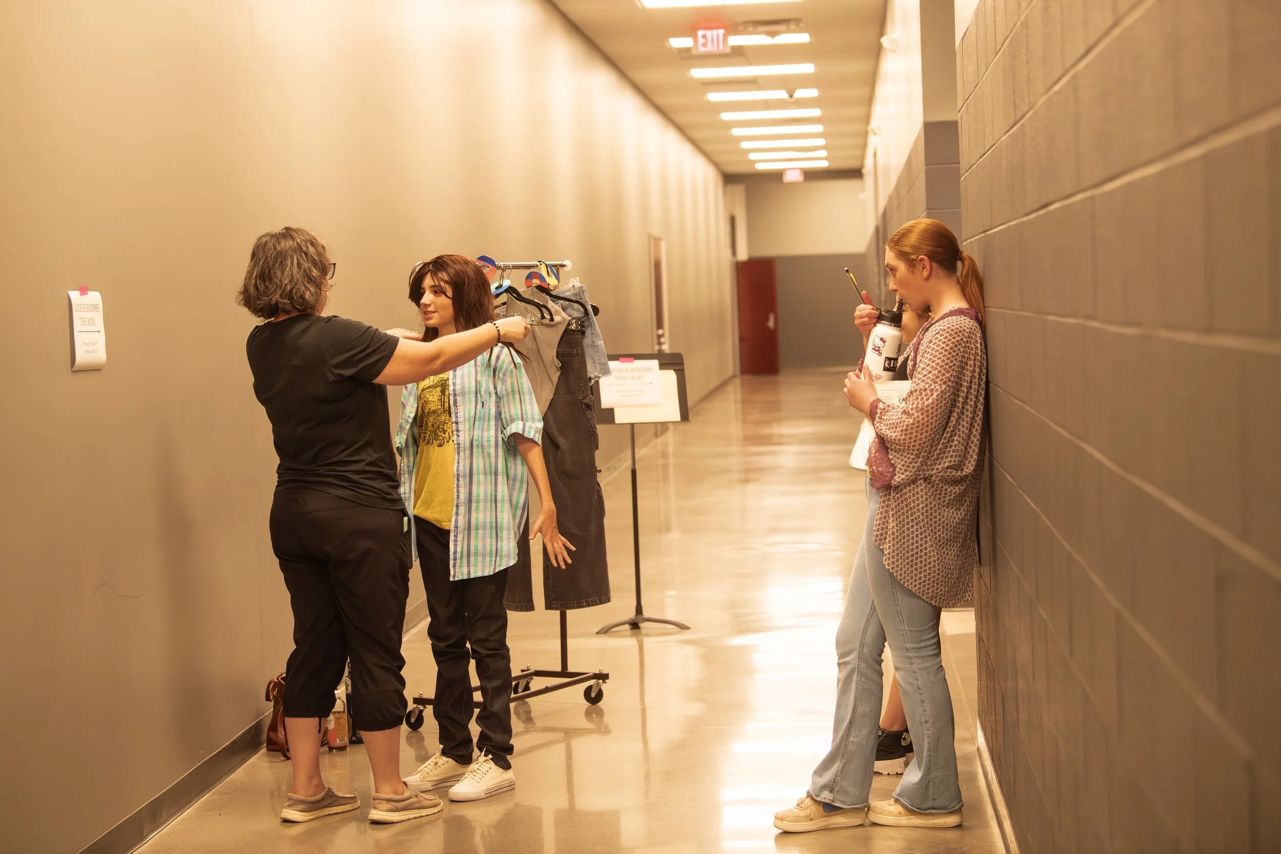 Three women in a hallway, one is a stylist helping a young woman with a short haircut and colorful outfit, another woman is leaning against the wall drinking from a cup, while a person in the background is partially visible.