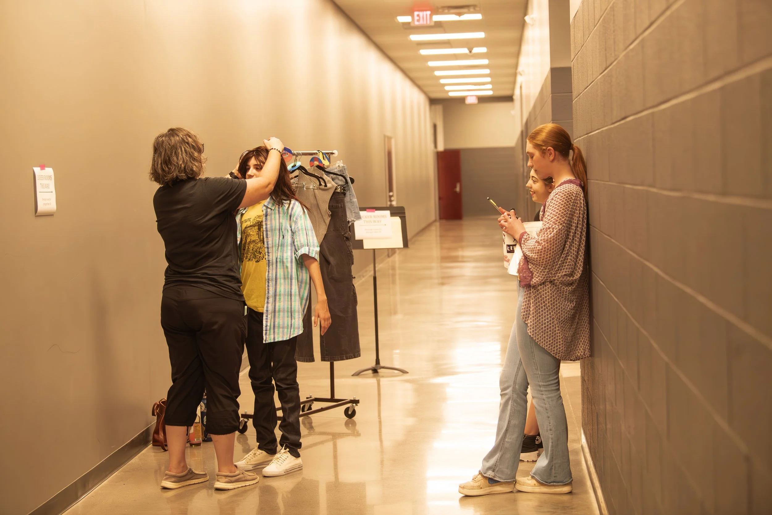People in a hallway at a costume fitting, with one person adjusting a costume on a model and others waiting while looking at their phones.