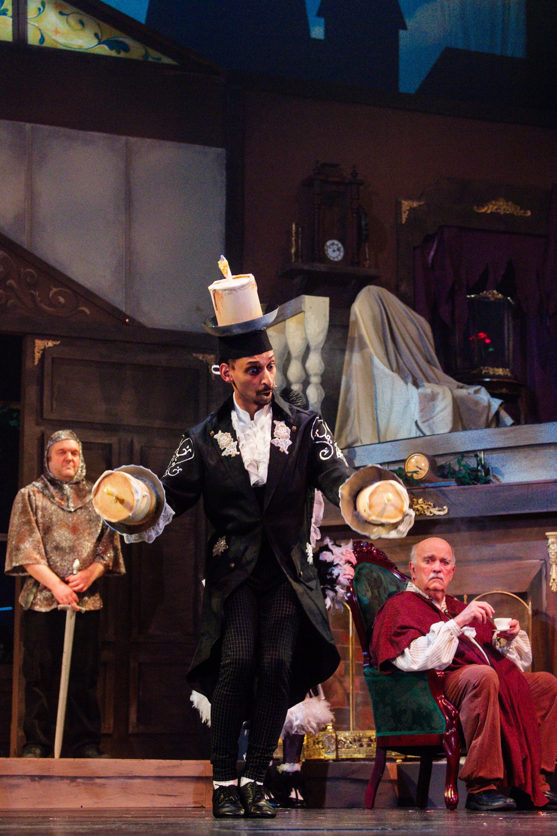 A theatrical stage scene with actors in costume. A man in the center is wearing a black coat with white embroidery, plaid pants, and a top hat with a candle on top, holding two hats. Behind him, an older man is seated in a green armchair, drinking from a cup, and another man stands in the background wearing a patterned shirt and a bonnet. The set depicts an elaborate interior with period furniture and decor.