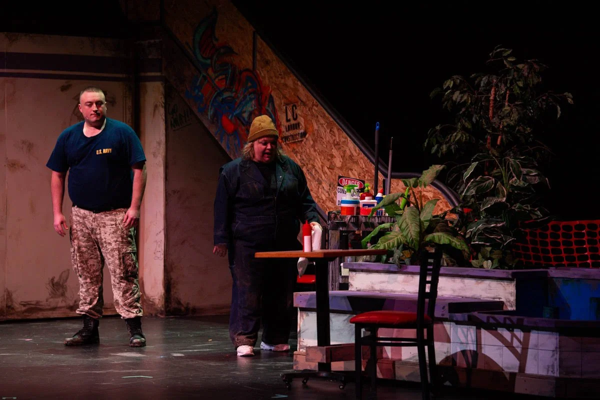 Two actors on stage, a man in U.S. Navy shirt and camo pants and a woman in dark work clothes, next to a table with various bottles and plants, in front of a graffiti wall.