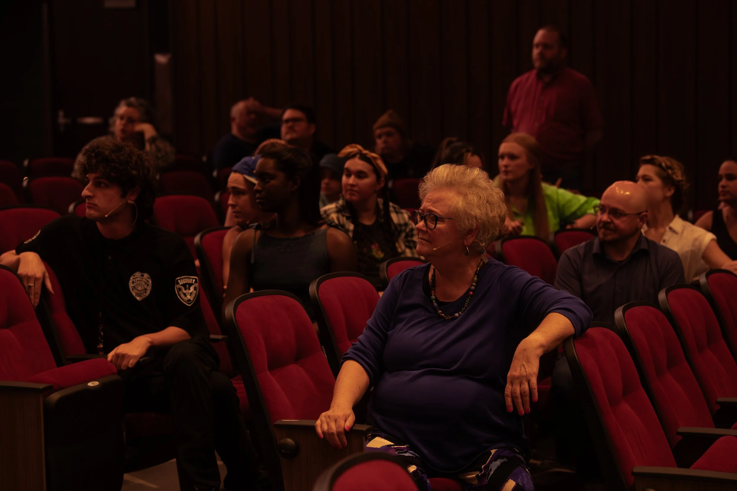 Audience seated in red theater seats during a presentation, some with microphones, in a dimly lit auditorium.