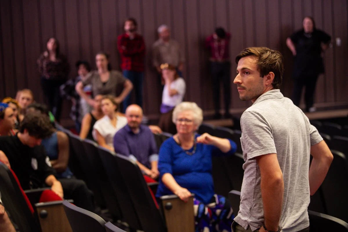 Young man standing in front of seated audience in a theater or auditorium.