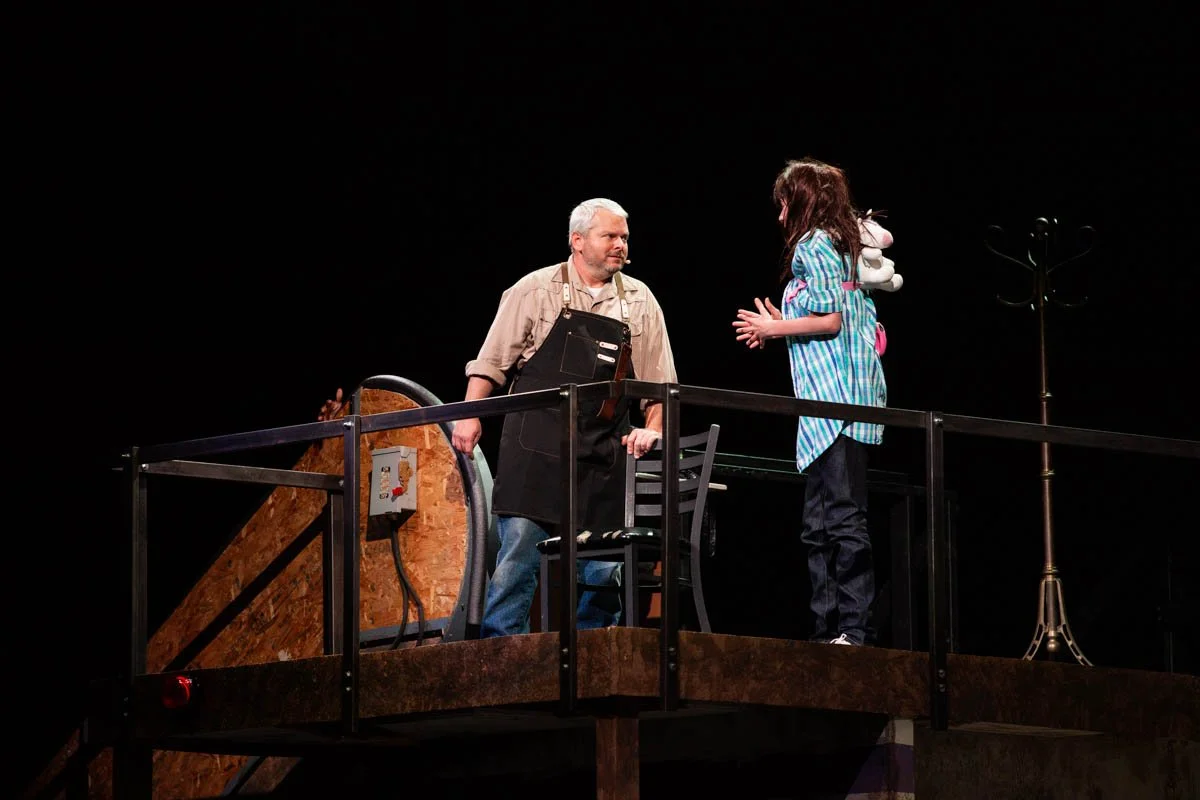 A theatrical scene with a man and a young girl on a stage set, possibly indicating a home or casual setting, with furniture and a coat rack present.