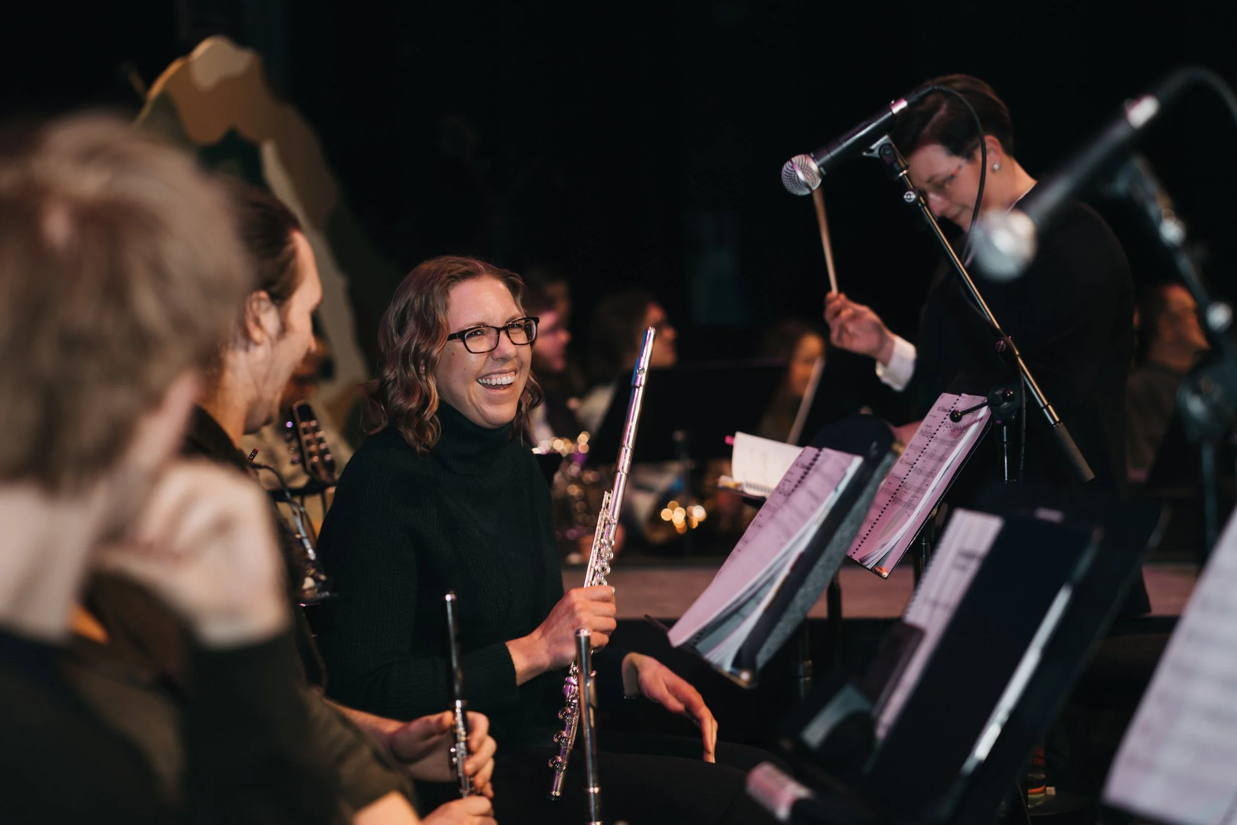 Orchestra musicians rehearsing, with a woman smiling and holding a flute.