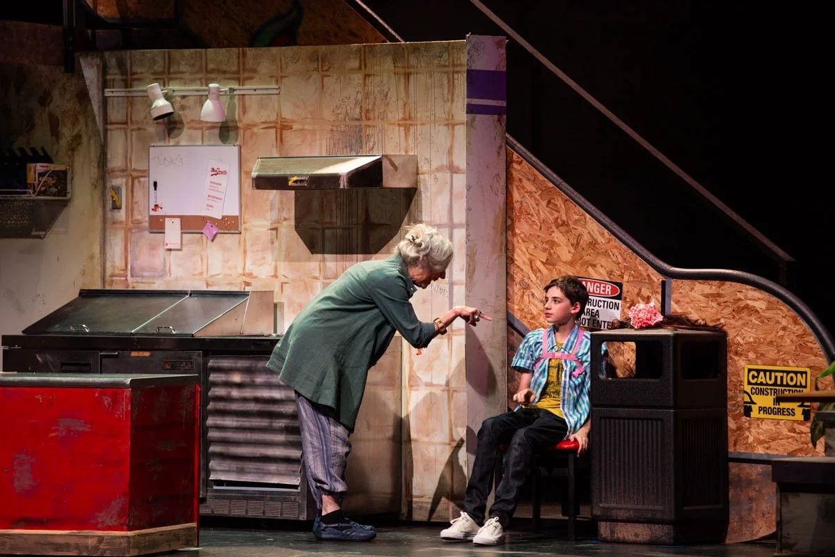 A woman with gray hair and a woman with dark hair are in a scene that looks like a kitchen set. The gray-haired woman is leaning forward, pointing and talking to the younger woman, who is sitting on a black chair. The scene has construction signs, a 