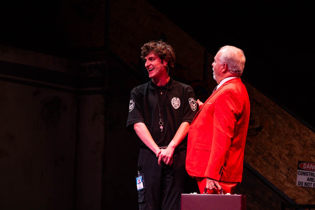 Two men in conversation on a stage with one dressed as a police officer and the other in a bright red suit. The police officer is smiling and wearing a uniform with patches, while the man in red holds a briefcase.