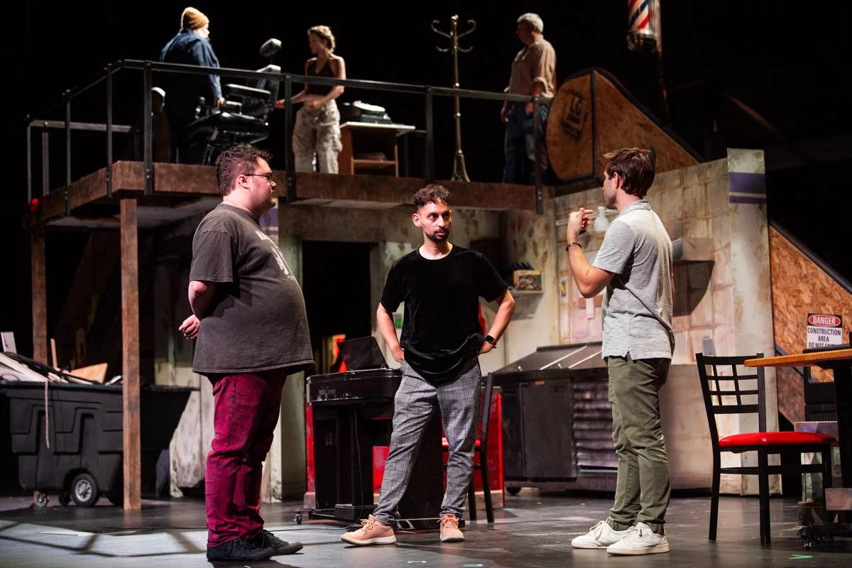 Three men standing in a theater rehearsal space, talking to each other, with a set behind them. Two women are on a balcony stage in the background.