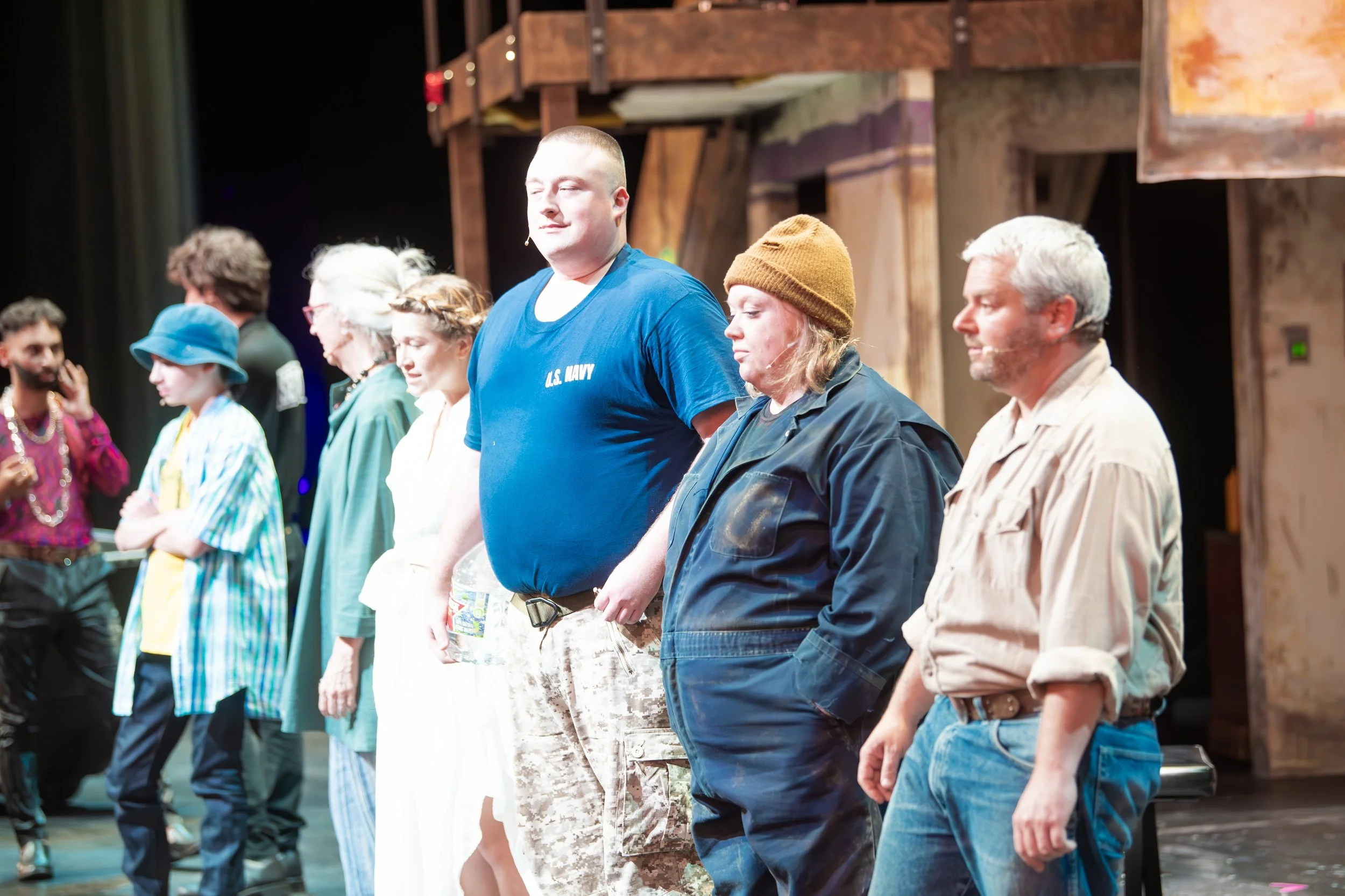 Line of diverse actors in costume on stage, participating in a theatrical production, with some taking a bow or listening.