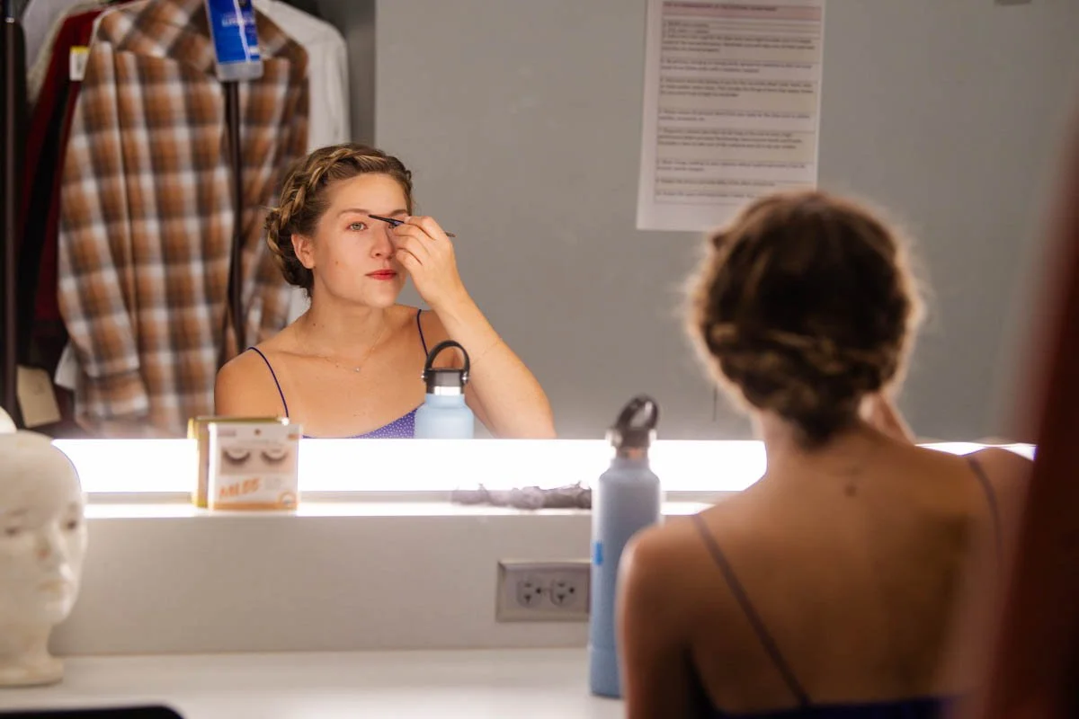 A woman with short hair and a braid, wearing a blue dress, is looking into a mirror and touching her eye. She is sitting in a dressing room or backstage area, with makeup or costume items visible, including a mannequin head with makeup on the counter