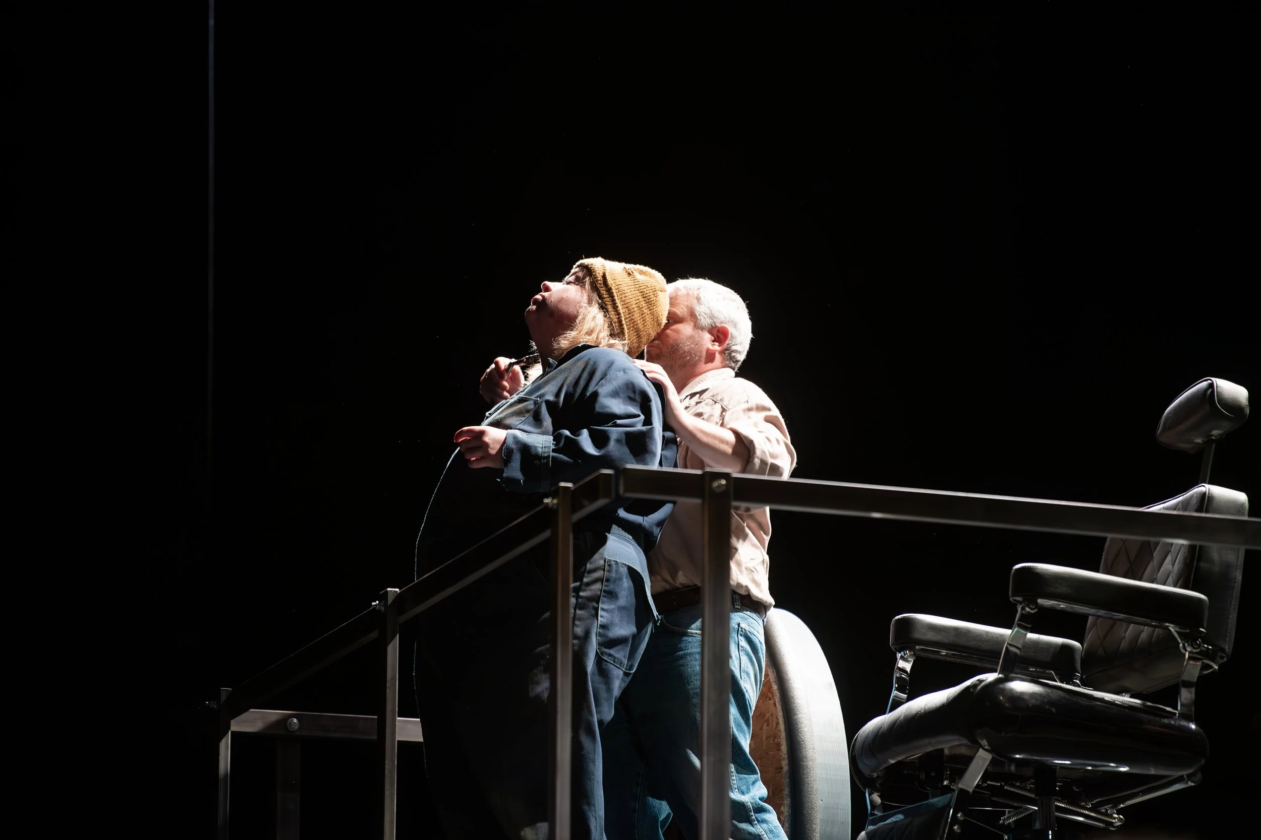 Two actors on stage, one kissing the other's cheek, with a black background and a stage railing in front.