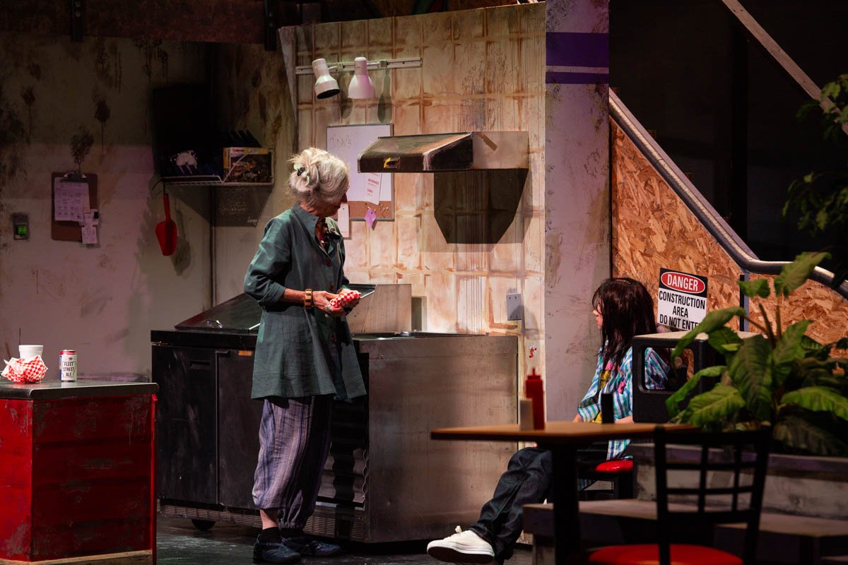 An elderly woman with gray hair and a man with long dark hair are in a kitchen or restaurant setting, with the woman inspecting a food container and the man sitting at a table.