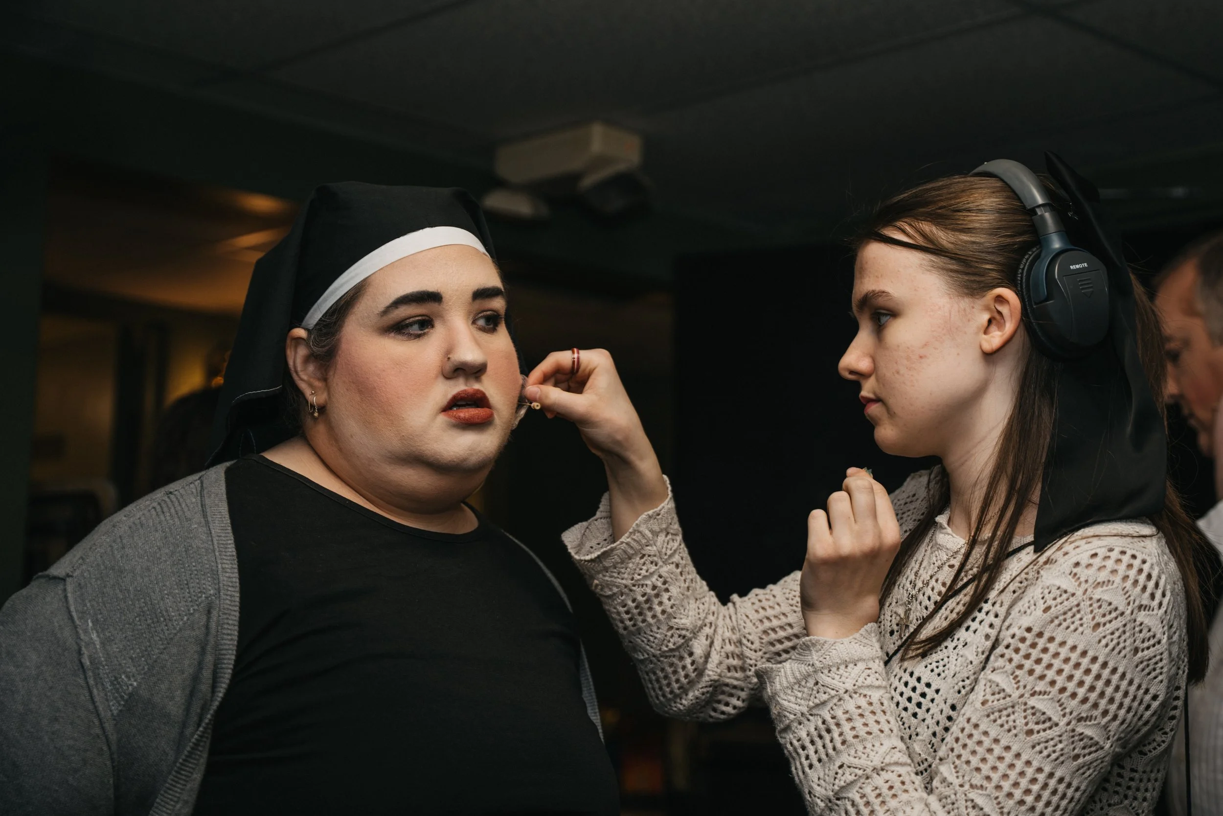 Makeup artist applying lipstick to woman with nun's habit in a dimly lit indoor setting