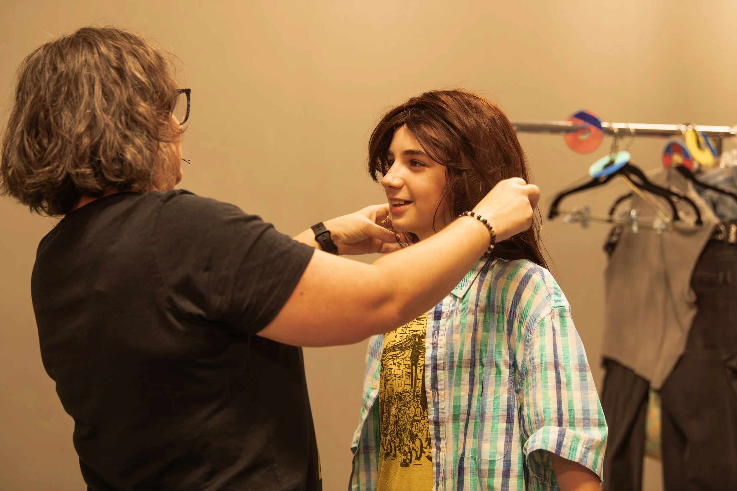 A woman with shoulder-length brown hair adjusting a young girl's hair in a dressing room or backstage area. The girl has medium-length red hair and is wearing a plaid shirt over a yellow t-shirt with a graphic design. Behind them is a clothing rack w