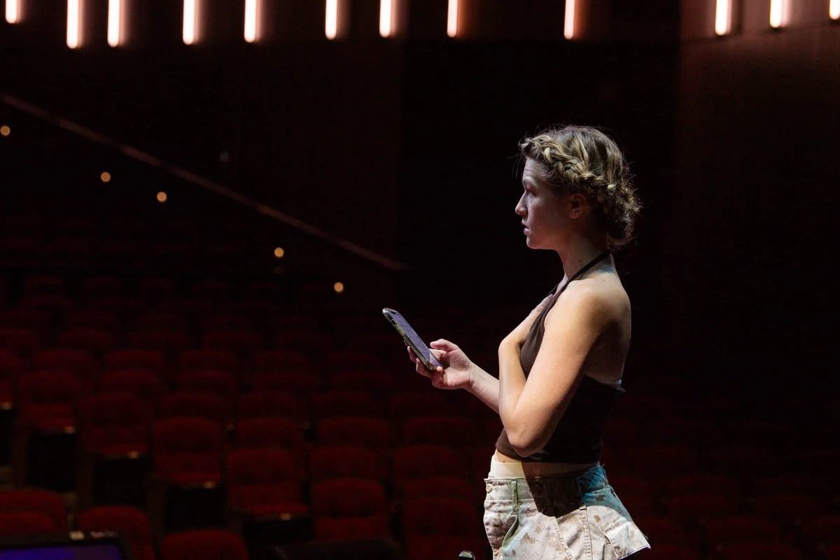 Young woman with braided hair standing alone in an empty theater, looking at her phone.