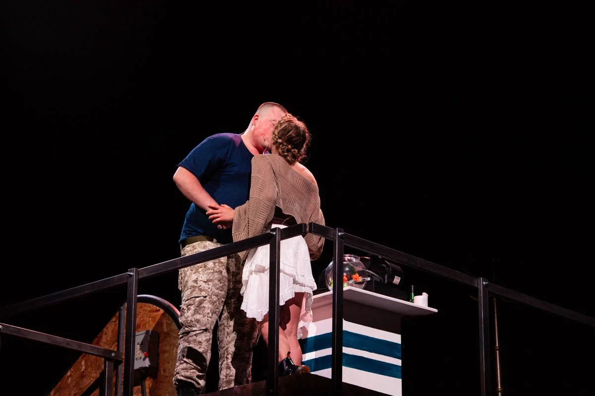 A man in military camouflage pants and a woman in a dress sharing a kiss on a stage with a dark background.