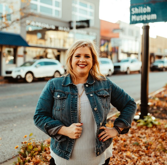 Smiling woman in denim jacket standing on street with cars and buildings in background