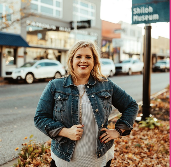 A smiling woman standing on a city sidewalk, with cars and shopfronts behind her, wearing a denim jacket and a white striped shirt.