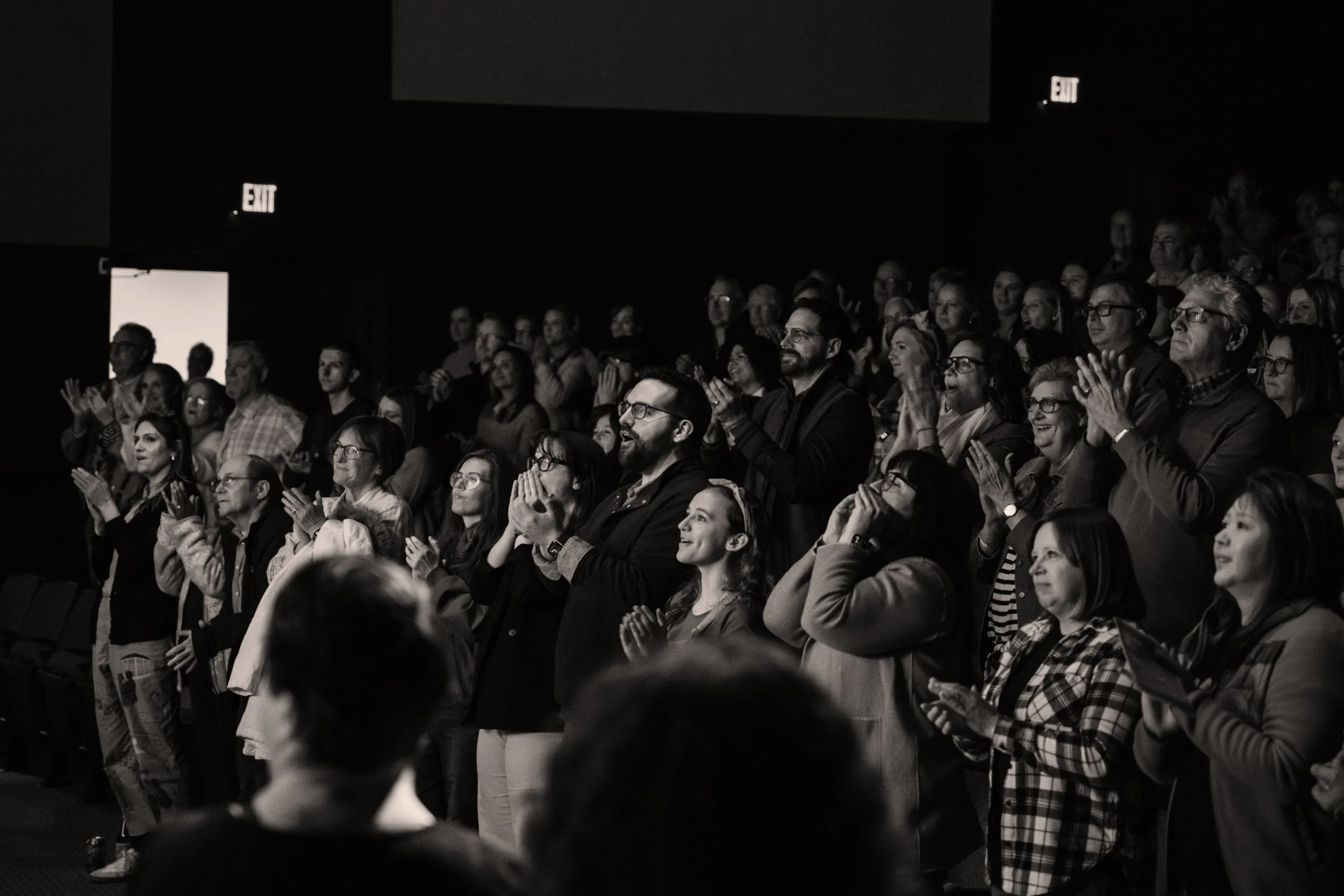 Audience standing, clapping, and smiling during a performance or event in a dimly lit theater.
