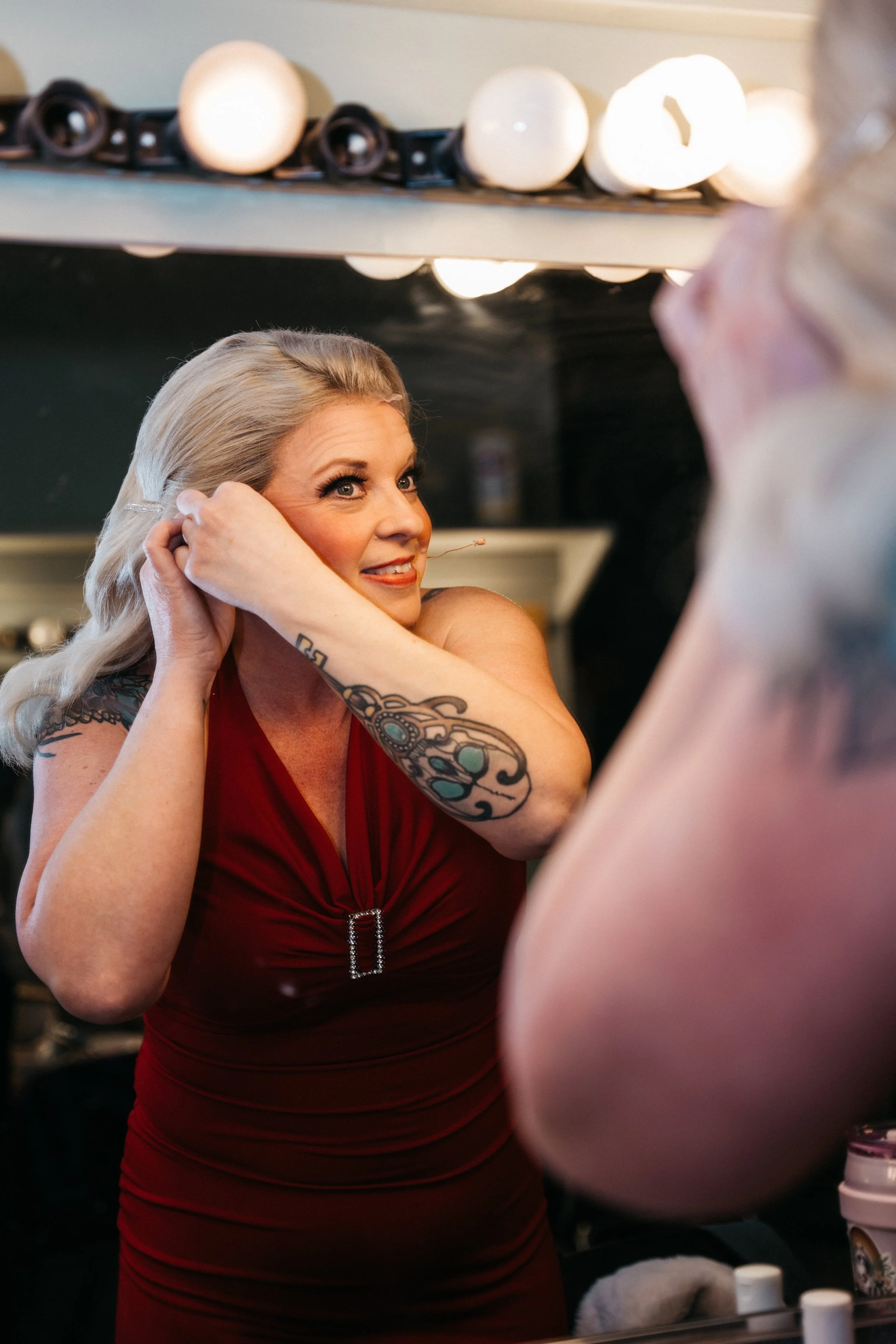 A woman in a red dress with tattoos is looking at herself in a mirror while putting on earrings, preparing for an event.