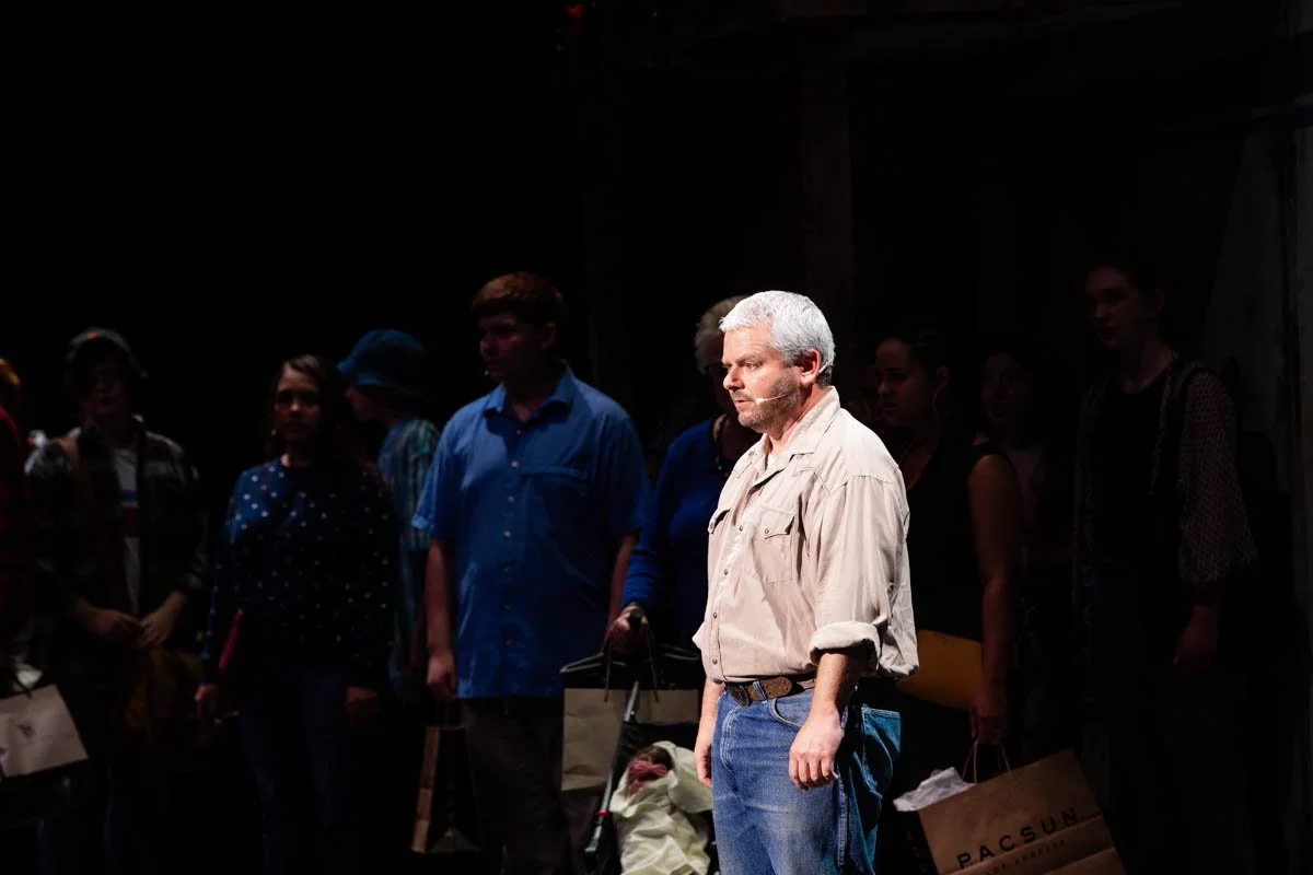 A man in a beige shirt and blue jeans stands alone in spotlight on stage, surrounded by a group of people mostly in shadow, holding shopping bags, with a dark background.