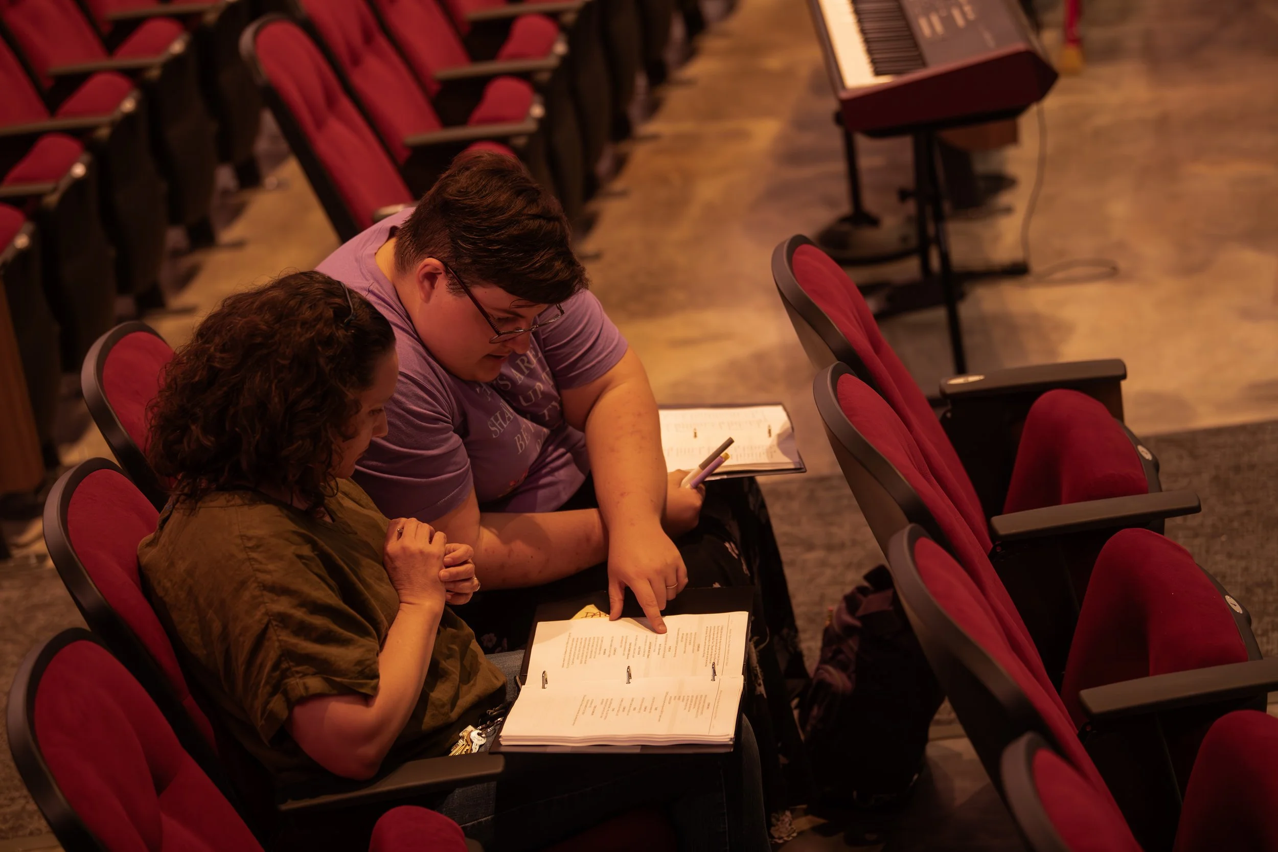 Two people sitting in a theater or auditorium, looking at open books and a cell phone, with empty red seats around them.