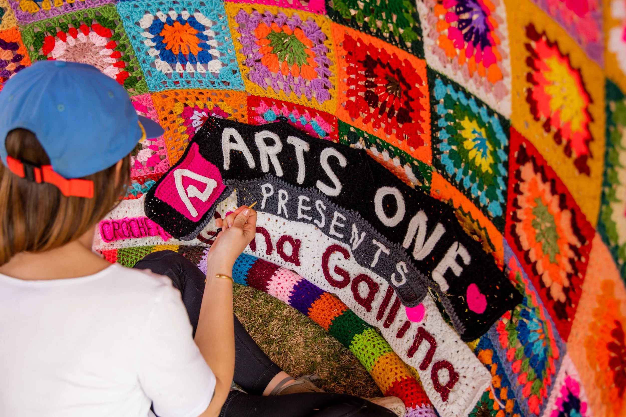 A woman with a blue cap working on a colorful crochet or knitted mural that features the text 'ARTS ON THE' and other words. The mural is made of various brightly colored squares and shapes.