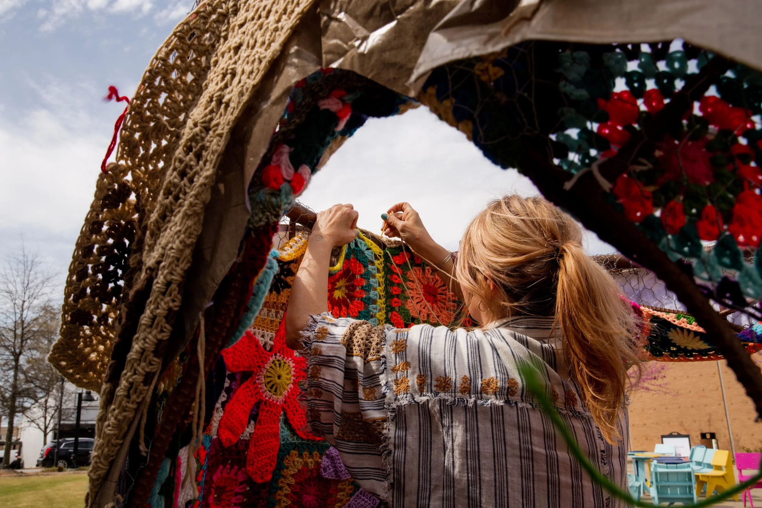 A woman with a ponytail knitting colorful granny squares of various patterns outside under a fabric structure, with a sky and some trees in the background.