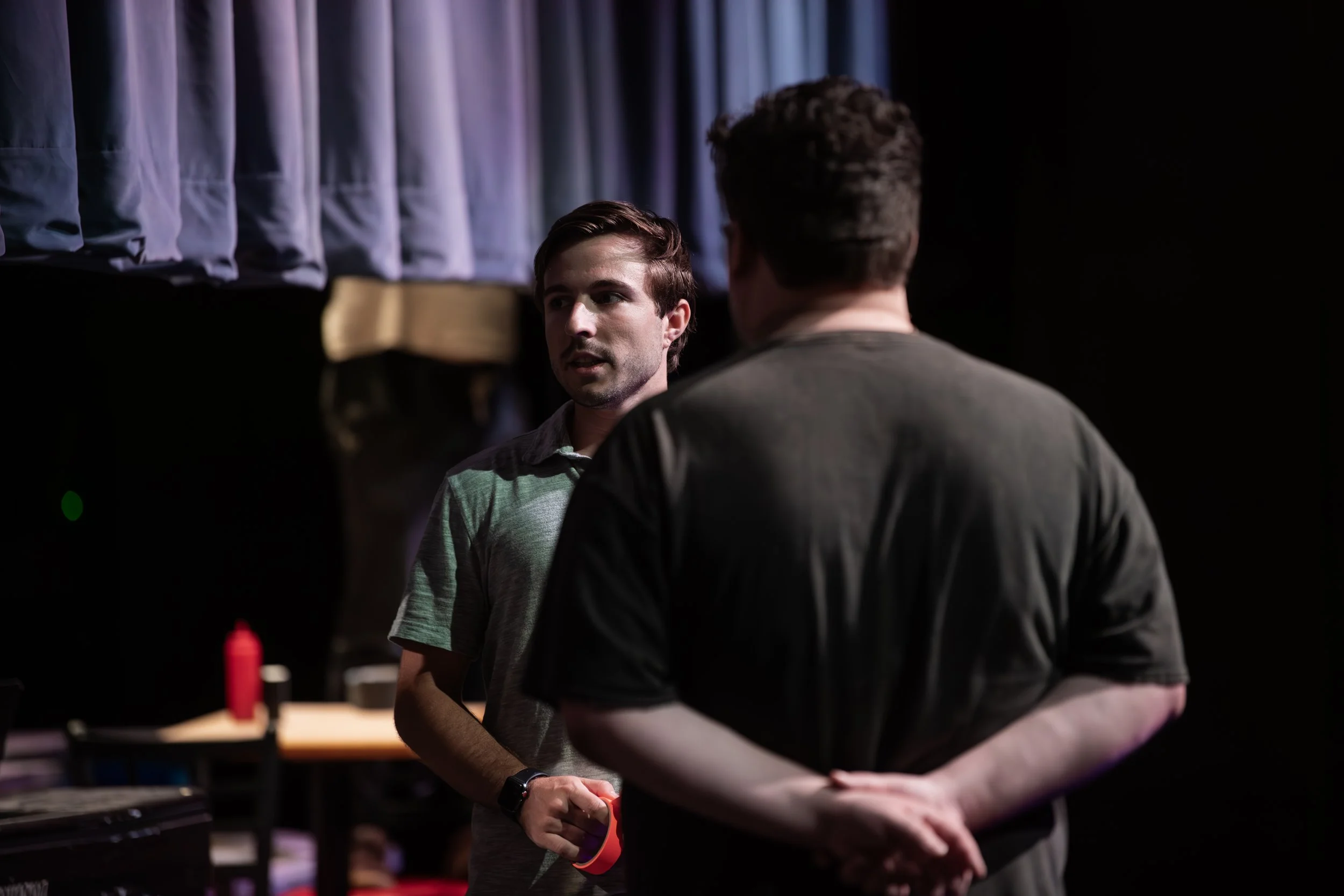 Two men standing and talking backstage with a dark background and a curtain.