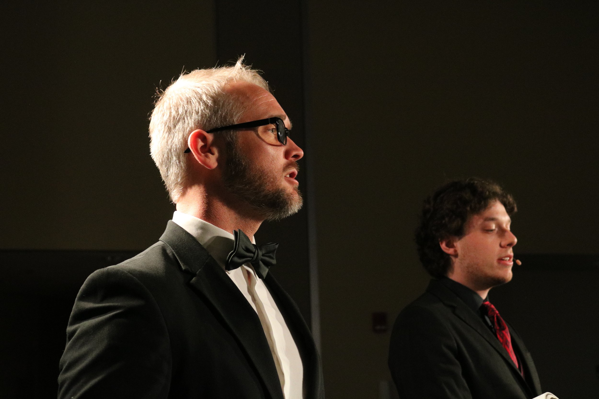 Two men in suits with ties, one with glasses and a beard, standing side by side in a dimly lit room, appearing to be at a formal event or presentation.