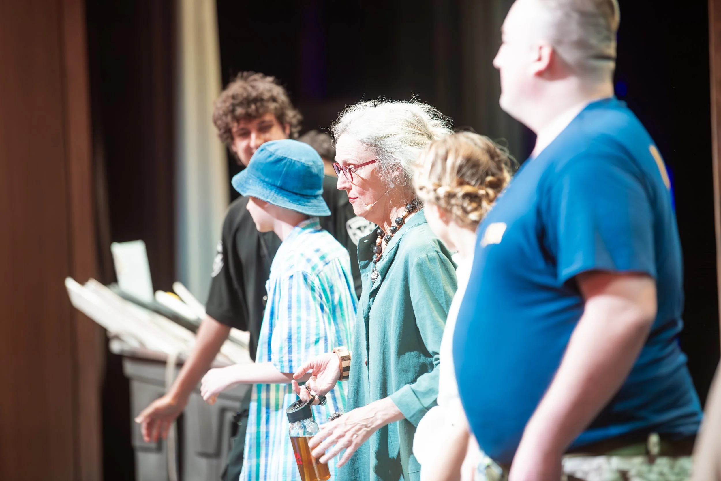 Group of people standing in a line, taking part in a standing activity, possibly on stage or in a workshop.