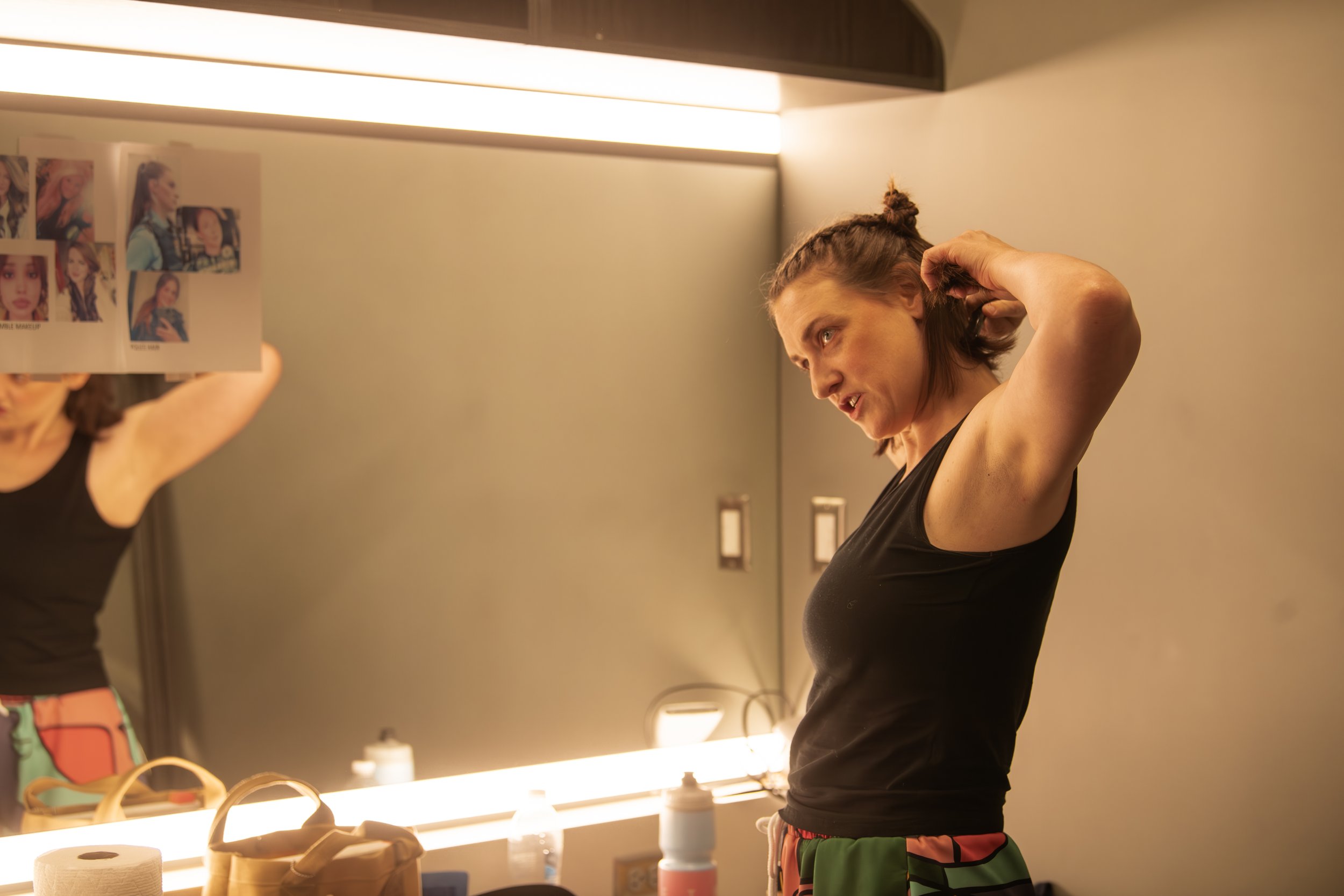 A woman with brown hair in a bun putting on makeup in front of a mirror in a dressing room. She is wearing a black sleeveless top and colorful patterned shorts, adjusting her hair.