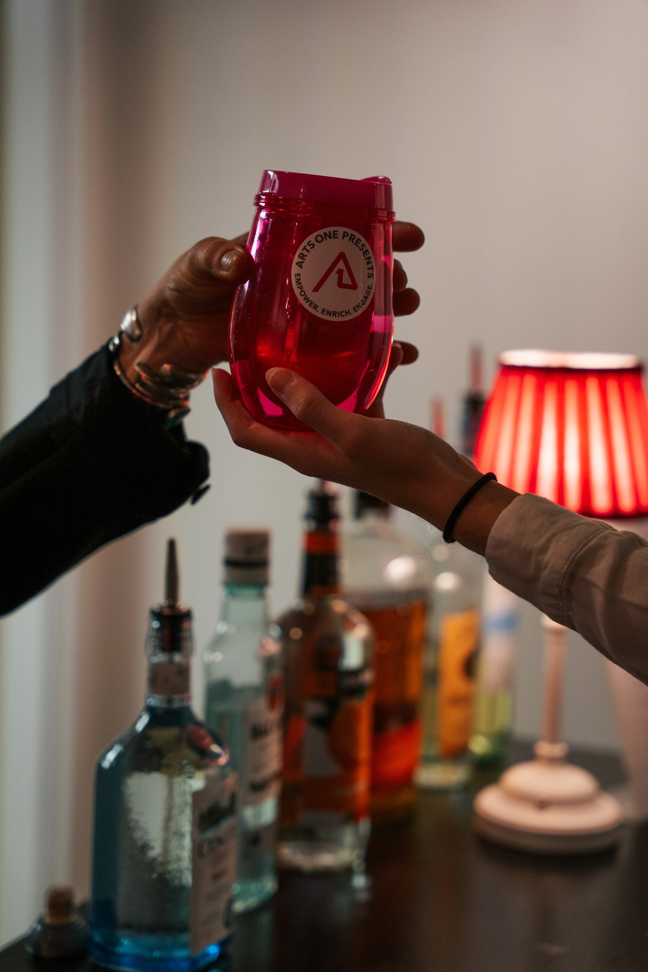 Two hands are clinking glasses, one hand with a red plastic cup and the other with a clear glass. The background features bottles of alcohol and a red-striped lamp.